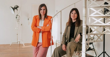 Zwei Frauen mit dunklen, schulterlangen Haaren. Links in orangen Hosenanzug gekleidet und stehend mit verschränkten Armren. Rechts auf weißer Wendeltreppe sitzend, in graubraunem Hosenanzug.