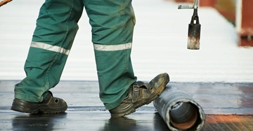 Person trägt grüne Arbeitskleidung und benutzt einen Gasbrenner um ein Bitumen-Dach zu schweissen, steht auf halb entrollter Bitumenrolle auf einem Dach im Freien