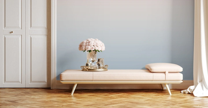 Eine beige Bank auf Holzfußboden darauf ein Tablett mit einer Vase mit Strauß aus rosa Rosen umgeben von einer blauen Wand und weißen Schränken links.