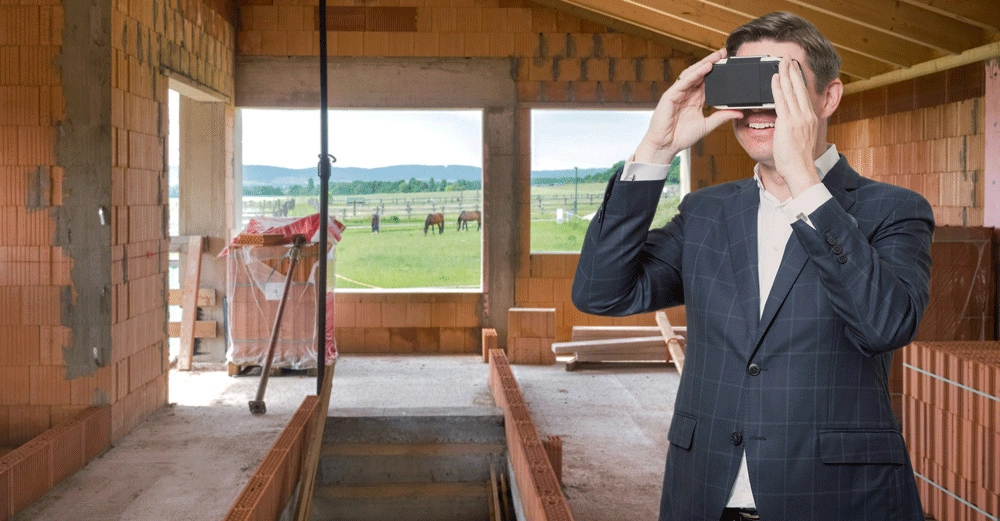 Ein Mann hält ein VR-Headset und lächelt, während er in einem Rohbau steht. Draußen vor dem Fenster sieht man grüne Wiesen und Pferde.