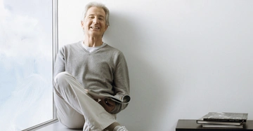 Ein Mann sitzt lächelnd auf einer Fensterbank hält eine Zeitschrift und trägt einen grauen Pullover und weiße Hosen. Neben ihm steht ein Tisch mit Büchern in einem hellen Zimmer.