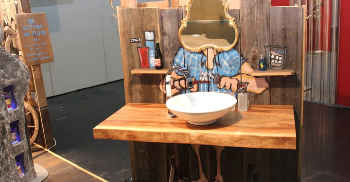 Galerie-Bild 7: Ein weißes Waschbecken mit einem Wasserhahn steht auf einem Holztisch in einer rustikalen Umgebung mit hölzernen Wänden. Ein Spiegel und bemalte Grafiken von Waschutensilien sind vorhanden. Text: NO SITTING ON FLOOR VIOLATORS WILL BE SHOOT SURVIVORS WILL BE SHOOT AGAIN STAMM DESIGN