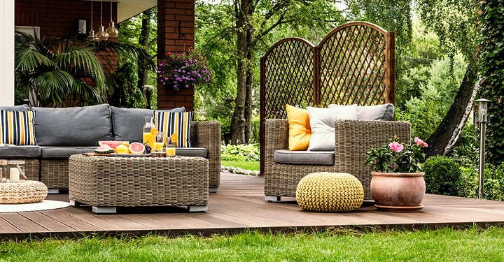 Geflechtmöbel und bunte Kissen stehen auf einer Holzterrasse. Ein Couchtisch mit Fruchtplatten und Getränken ist in der Mitte. Im Hintergrund Pflanzen und ein Garten mit Bäumen und Blumen.