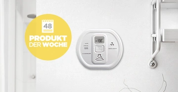 Ein Kohlenmonoxid-Melder hängt an einer weißen Wand in einem modernen, hellen Raum. Links ist ein gelber Kreis mit der Aufschrift Kalender 48 PRODUKT DER WOCHE.