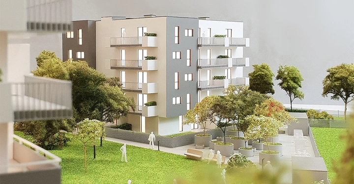 Modellgebäude mit mehreren Stockwerken und Balkonen steht umgeben von Bäumen und Rasenfläche. Stilisierte Figuren stehen und sitzen im Gartenbereich. Weitere Gebäude sind teilweise sichtbar im Hintergrund.