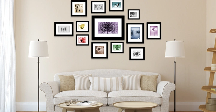 Drei Tische und Sofa stehen vor einer hellen Wand auf der Bilder in schwarzen Rahmen aufgehängt sind Zwei Stehlampen flankieren das Sofa und ein Regal steht rechts