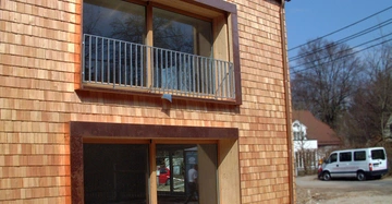 Holzverkleidetes zweistöckiges Haus mit großen Fenstern und einem kleinen Balkon im Obergeschoss in einem Wohngebiet mit geparktem weißen Kleinbus und kahlen Bäumen im Hintergrund.