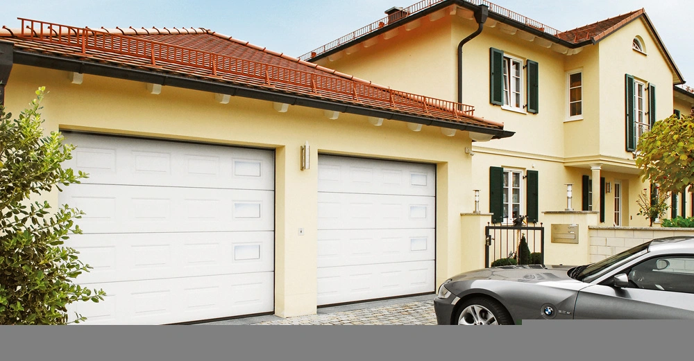 Ein graues Auto parkt vor einer großen gelben Villa mit roten Dachziegeln. Die Villa hat zwei weiße Garagentore und grüne Fensterverschlüsse. Ein Baum und Sträucher umrahmen die Szene.