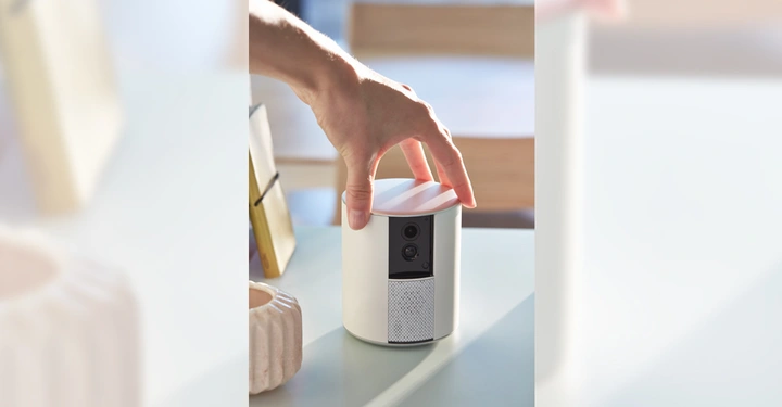 Hand hält eine zylindrische, weiße, elektronische Kamera auf einem Tisch in einem gut beleuchteten Raum mit Holzmöbeln.