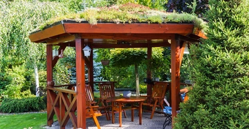 Pavillon aus tropischem Holz, gebeizt oder lasiert, mit begrüntem Dach. Darunter eine Sitzgruppe mit Sesseln und Tisch aus Holz. Geplasterter Boden, rundum Garten