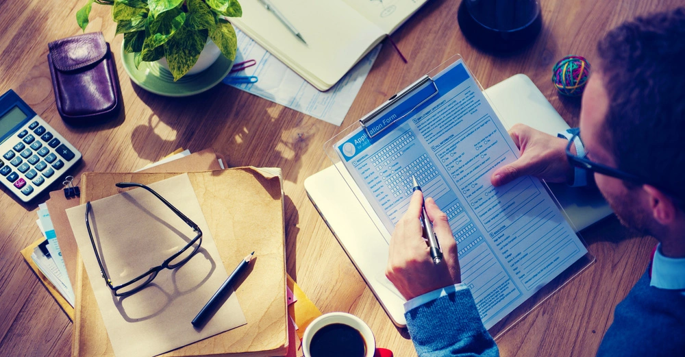 Mann füllt ein Formular aus und sitzt an einem Schreibtisch mit Unterlagen, Kaffeetasse, Brille, Taschenrechner, Notizbuch, Pflanzen und einem Bürozubehör.