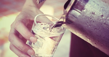 Eine Hand hält ein Glas, in das Wasser aus einem Metallkrug gegossen wird; im Hintergrund unscharfe Außenbereich.