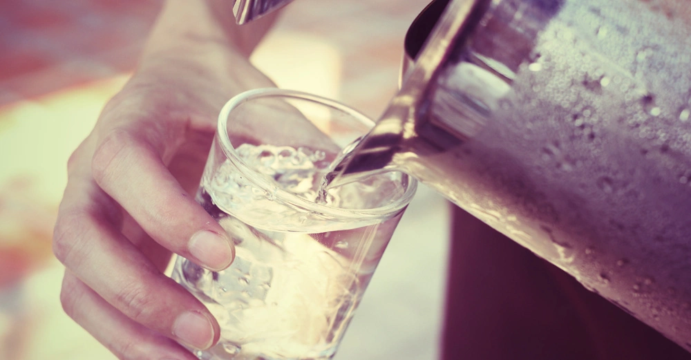 Eine Hand hält ein Glas, in das Wasser aus einem Metallkrug gegossen wird; im Hintergrund unscharfe Außenbereich.