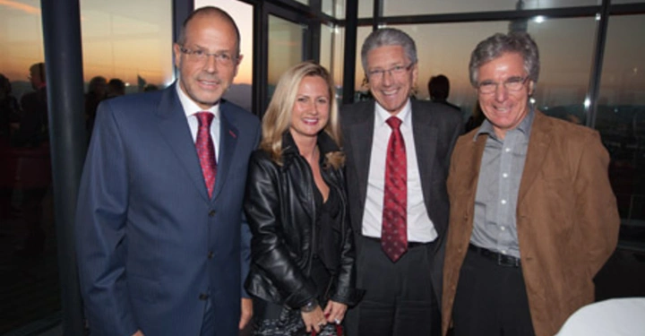 Galerie-Bild 60: Vier Personen stehen und lächeln in die Kamera Kontext ist ein Raum mit großen Fenstern während des Sonnenuntergangs im Hintergrund Die Personen tragen formelle Kleidung und stehen nah beieinander