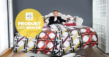Eine Person sitzt in einem weiß verzierten Bett mit bunter Bettwäsche in einem modernen Schlafzimmer. Text: "45 PRODUKT DER WOCHE".