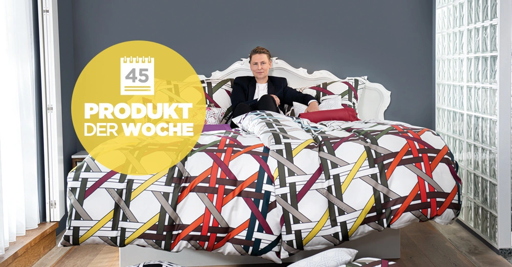 Eine Person sitzt in einem weiß verzierten Bett mit bunter Bettwäsche in einem modernen Schlafzimmer. Text: "45 PRODUKT DER WOCHE".