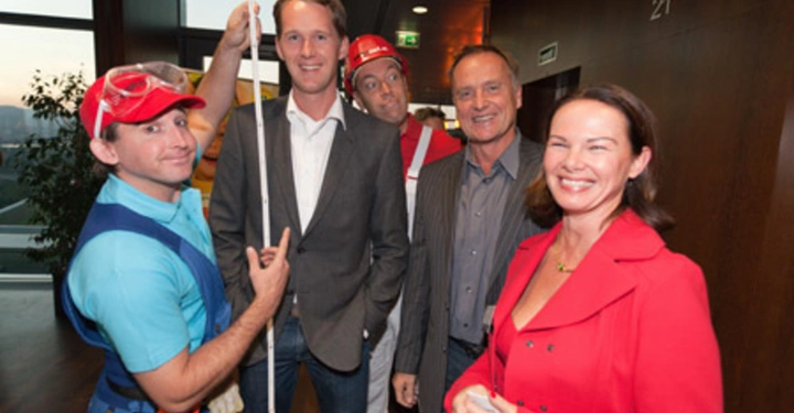 Galerie-Bild 51: Mehrere Personen stehen in einem Innenraum und lachen der im Vordergrund ein Mann in einem blauen Hemd hält einen langen weißen Stab die Frauen und Männer sind formell gekleidet. Eine Tür mit der Nummer 21 ist sichtbar.