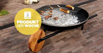 Großer runder Grill steht auf gepflastertem Boden grillt Mais und Fleischspieße. Links ein gelber Kreis mit einem Symbol eines Kalenders und Text: 2 PRODUKT DER WOCHE