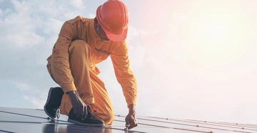 Arbeiter kniet auf einem Dach und installiert Sonnenkollektoren an einem sonnigen Tag