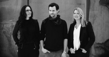 Drei Personen stehen nebeneinander und lächeln freundlich in die Kamera. Sie tragen formelle Kleidung. Der Hintergrund ist eine Betonwand.