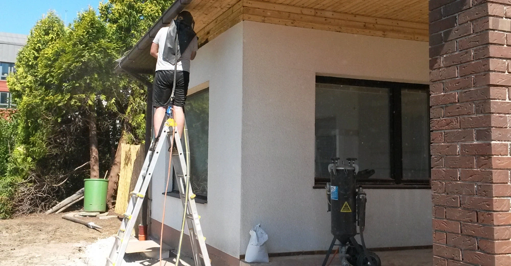 Eine Person steht auf einer Leiter und arbeitet an der Fassade eines Hauses mit weißer Wand und Holzverkleidung. Im Hintergrund befinden sich Bäume und Bauzubehör.