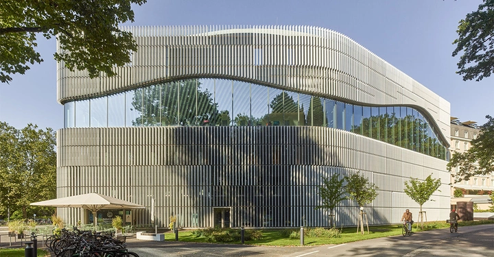 Ein modernes Gebäude mit wellenförmiger Fassade steht in grüner Umgebung Bäume und Fahrradständer vor dem Bau Radfahrer fahren auf einem Weg am Gebäude entlang.