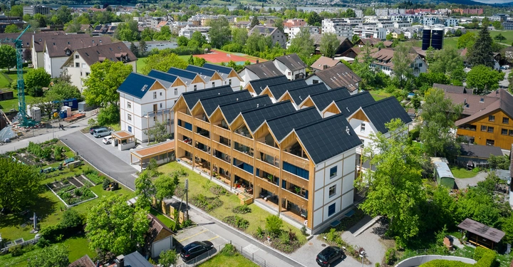 Mehrstöckige Gebäude mit Solardächern stehen in einer grünen, ländlichen Umgebung. Einige parkende Autos sind auf den Straßen sichtbar, und im Hintergrund befinden sich andere Häuser und Gebäude.