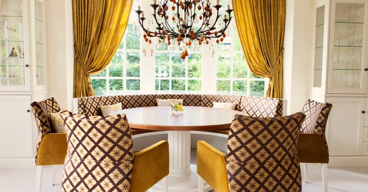 Runder Holztisch mit gemusterten Stühlen steht unter einem Kronleuchter in einem eleganten Esszimmer eingerahmt von großen Fenstern mit goldenen Vorhängen und weißen Vitrinen auf beiden Seiten.