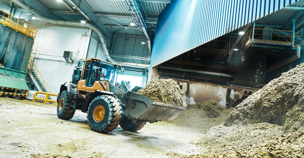 Ein Bagger hebt eine Schaufel voll Sand in einer großen, gut beleuchteten Industriehalle mit Metallstrukturen und Maschinen im Hintergrund