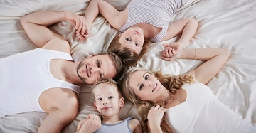 Vier Personen liegen auf dem Bett Hand in Hand und schauen zur Kamera sie tragen weiße und graue Kleidung das Bett hat weiße Laken.