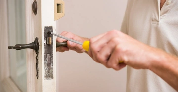 Person montiert mit einem Schraubendreher ein Türschloss in einer weißen Tür in einem Innenraum.