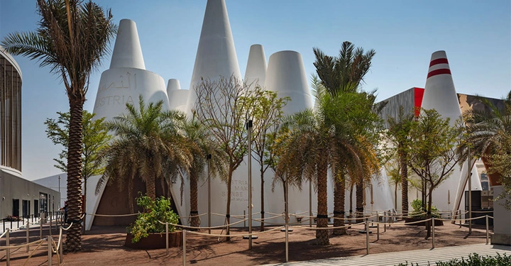 Galerie-Bild 1: Weiße kegelförmige Gebäude stehen zwischen mehreren Palmen und Bäumen an einem eingezäunten Weg mit schattigem Boden. Text auf den Gebäuden: Austria AMA MAK SENSE.