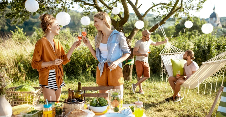 Gartenparty: 2 junge Frauen in sommerlichen Outfits stehen vor vollem Tisch und stoßen mit einem Weinglas an, im Hintergrund sitzt ein Mann in der Hängematte und unterhält sich mit einem anderen jungen Mann, der auf Holzschemel sitzt.