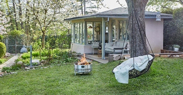Hängesessel schwingt an einem Baum im blühenden Garten vor einem gemütlichen Haus Zimmer mit Glasfenstern Feuerstelle brennt davor Teppich liegt im Sessel Laternen hängen umher heller Tag.