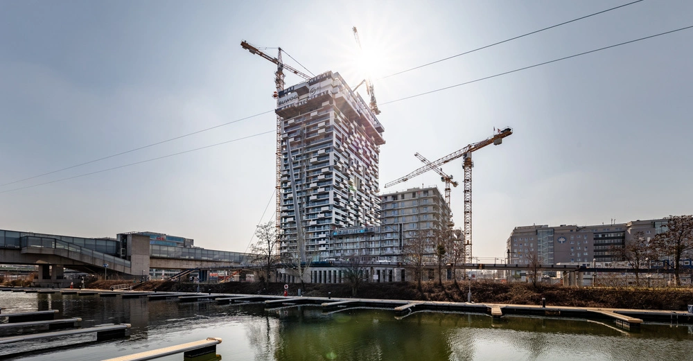 Ein im Bau befindlicher Wolkenkratzer mit Kränen und Baugerüsten umgeben von anderen Gebäuden am Flussufer bei Sonnenschein. Beschilderung zeigt die Aufschrift BUWOG MAR.