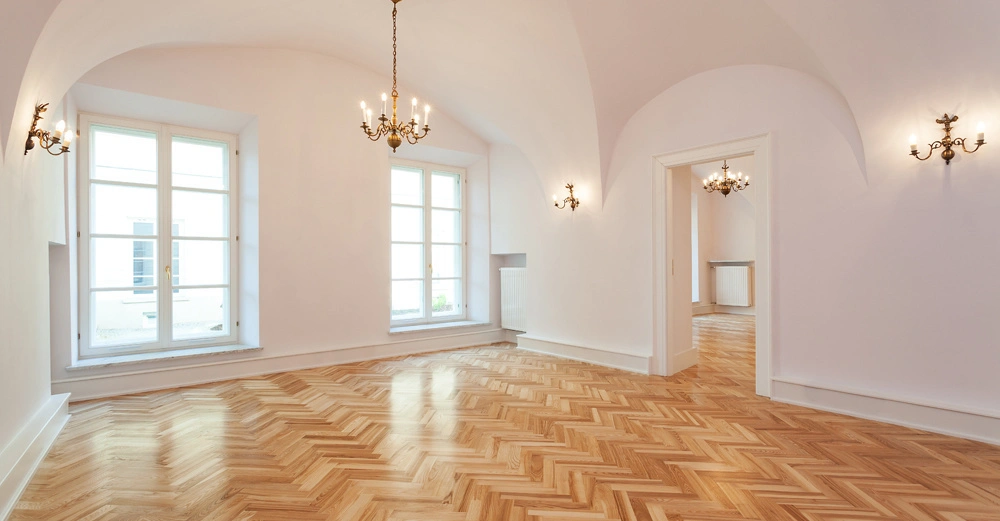 Ein leerer Raum mit Parkettboden und Kronleuchtern steht in einem weißen Interieur mit gewölbter Decke und großen Fenstern gegenüber einer offenen Tür zu einem anderen Raum