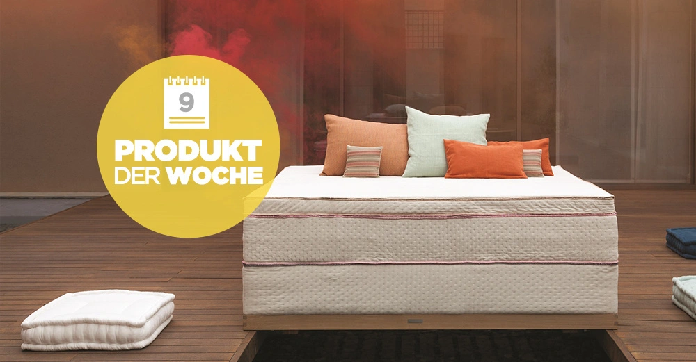 Bett mit weißen und bunten Kissen steht auf einer Holzterrasse vor einer Glaswand in einem modernen, hellen Ambiente. Ein großes Schild rechts zeigt: 9 PRODUKT DER WOCHE.