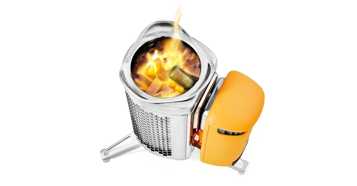 Metallischer Campingkocher mit brennendem Holz, ausgestattet mit einem orangefarbenen Anbaugerät, auf drei klappbaren Beinen in einer weißen Umgebung.