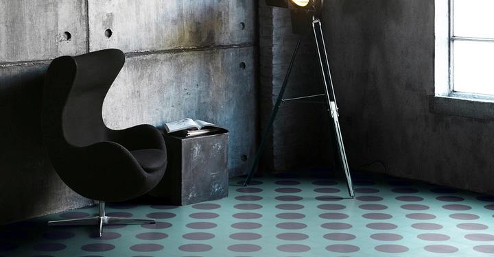 Galerie-Bild 8: Ein schwarzer Sessel steht neben einem Tisch mit einem offenen Buch auf einem gefliesten Boden mit großen Punkten in einem industriellen Raum mit Betonwänden und einem Stehstrahler.