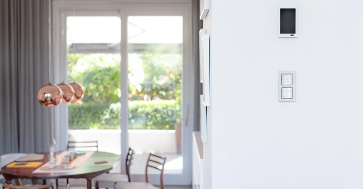 Ein moderner Raum mit einem Holztisch an dem Stühle stehen und darüber drei hängende Lampen. Im Hintergrund eine Glastür mit Blick auf einen grünen Garten.