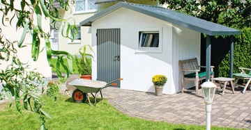 Ein Schuppen steht auf einer gepflasterten Terrasse Ein Schubkarren neben einem Blumentopf mit gelben Blumen Ziegelhäuser und Bäume im Hintergrund Ein Holzbank und Stühle unter einem Dachvorsprung