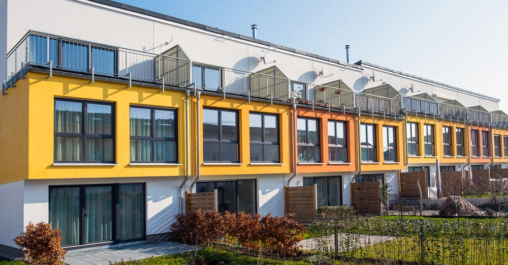 Mehrfamilienhaus mit bunten Fassaden im Gelb- und Orangebereich hat Balkone im Obergeschoss. Die Grünfläche davor ist gepflegt mit Büschen und einem kleinen Zaun. Klarer Himmel im Hintergrund.