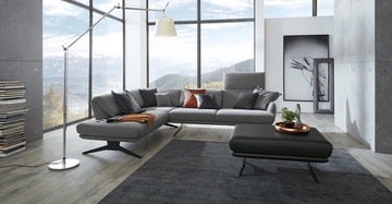 Großes, graues Sofa mit Kissen steht vor raumhohen Fenstern, mit Aussicht auf Berge; moderne Lampe, Bücherstapel, schwarzer Tisch, Pflanze und Kunstwerk im Hintergrund; Holzboden.