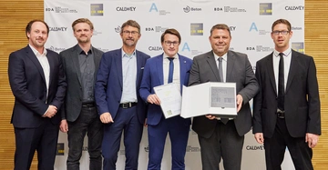 Sechs Männer tragen Anzüge und lächeln in die Kamera. Zwei halten Zertifikate und einen Preis. Sie stehen vor einer Wand mit Logos in einem gut beleuchteten Raum.