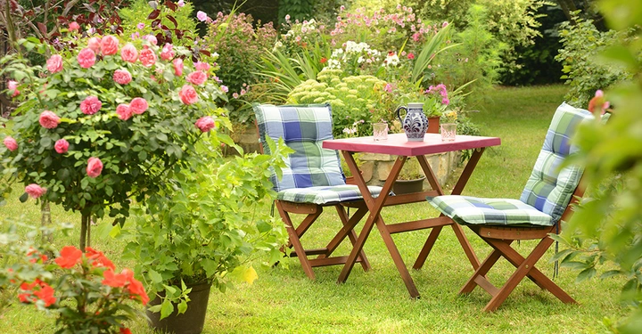 Holztisch mit Krug und Gläsern steht im Garten zusammen mit zwei gepolsterten Holzstühlen umgeben von blühenden Rosen und anderen Pflanzen.