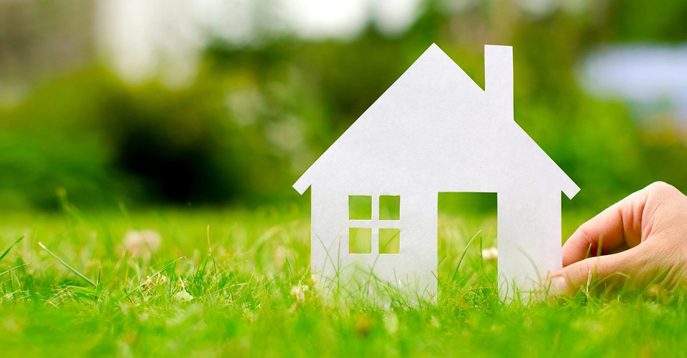 Papierhaus wird von einer Hand gehalten Wiese und Bäume im Hintergrund.