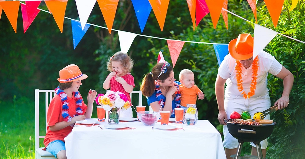Familie, ein Junge, ein kleineres Mädchen und ein Baby in Mamas Arm sitzen rund um Gartentisch, Vater steht am Grill und wendet Paprika. Partydeko in Orange, Blau und Weiß. Glückliche Familie