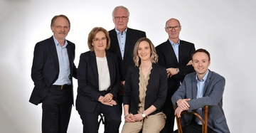 Sechs Personen posieren in formeller Kleidung vor einem hellen Hintergrund Drei stehen und drei sitzen auf Stühlen Die Gruppe lächelt und schaut in die Kamera