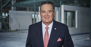 Ein Mann in einem dunkelblauen Anzug mit roter Krawatte lächelt in die Kamera in einer großen industriellen Halle mit sichtbaren Metallstrukturen im Hintergrund stehend.