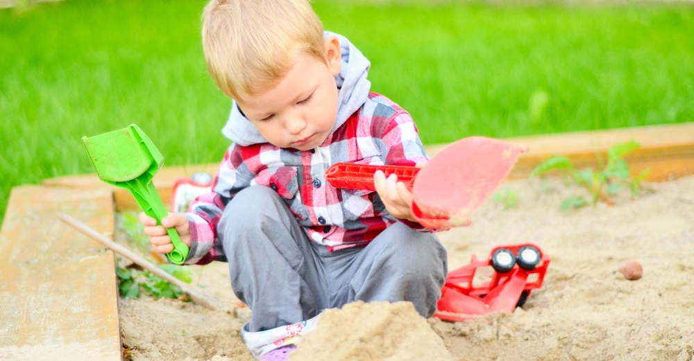 Kleinkind spielt im Sandkasten mit einer grünen Schaufel in der linken Hand und einer roten Schaufel in der rechten Hand umgeben von grünem Gras und Spielzeuglastwagen im Sand.